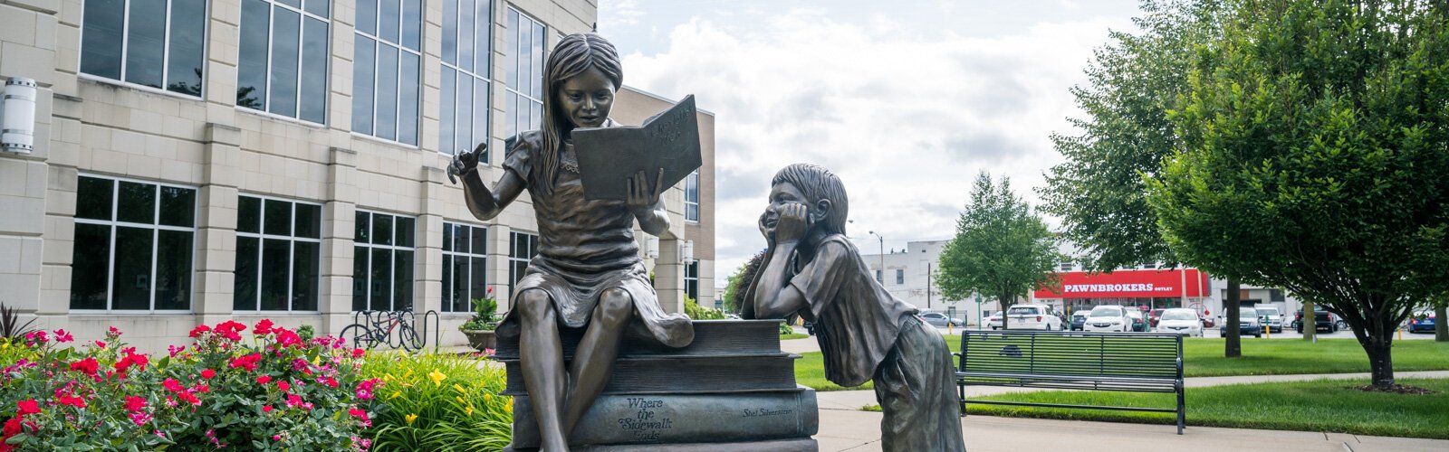 A playful statue of two children and a giant stack of books sits outside the main library, the Alice and Jack Wirt Public Library.