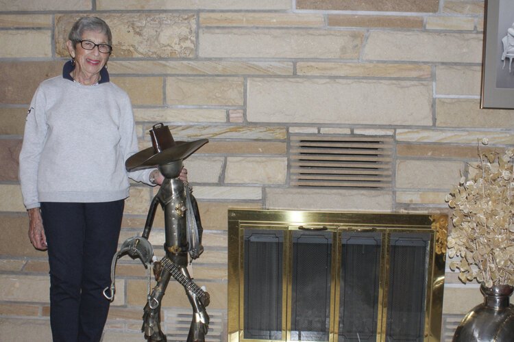 Rose Wunderbaum Traines stands with one of her metal sculptures that is prominent in her home – a cowboy that features a hat made from a cowbell, chaps crafted from shoe horns, and boots created out of hooks from old logging chain.
