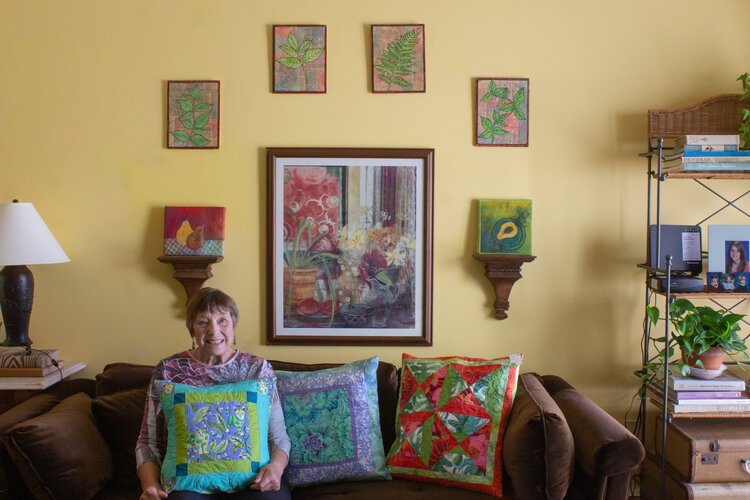 Virginia “Ginny” Crandall sits in her home, surrounded by pillows, wall hangings, and other fabric creations that she has made.