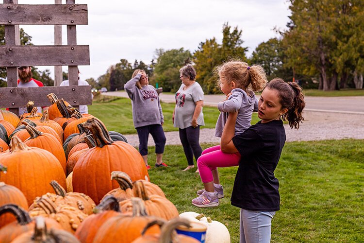 Maeve hoists Hattie Meyer, 3, onto the pumpkin display so she can choose the pumpkin she wants. Callie Meyer, 38, of Mount Pleasant, speaks to Becky while her daughter picks out her pumpkin for the season. 