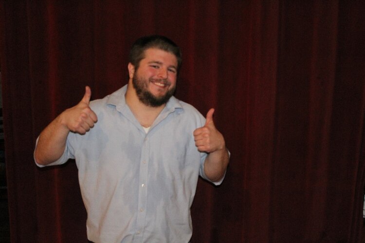 Jon Cauchi uses humor to raise awareness about living with a disaiblity. In this photo, Jon Cauchi is wearing a short sleeve light blue shirt. He is standing, holding his two thumbs up and smiling. 