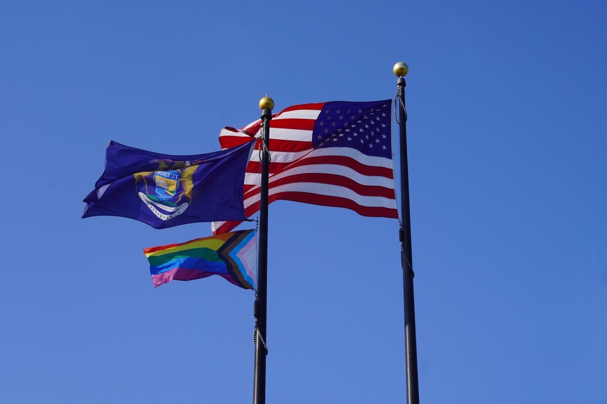 The US, Michigan State, and Progress Pride flags fly in the sky over City Hall.