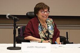 A photograph of Judith Heumann. She is sitting in her power chair behind table covered with a white table cloth. There is a microphone and a name placard in the foreground. 