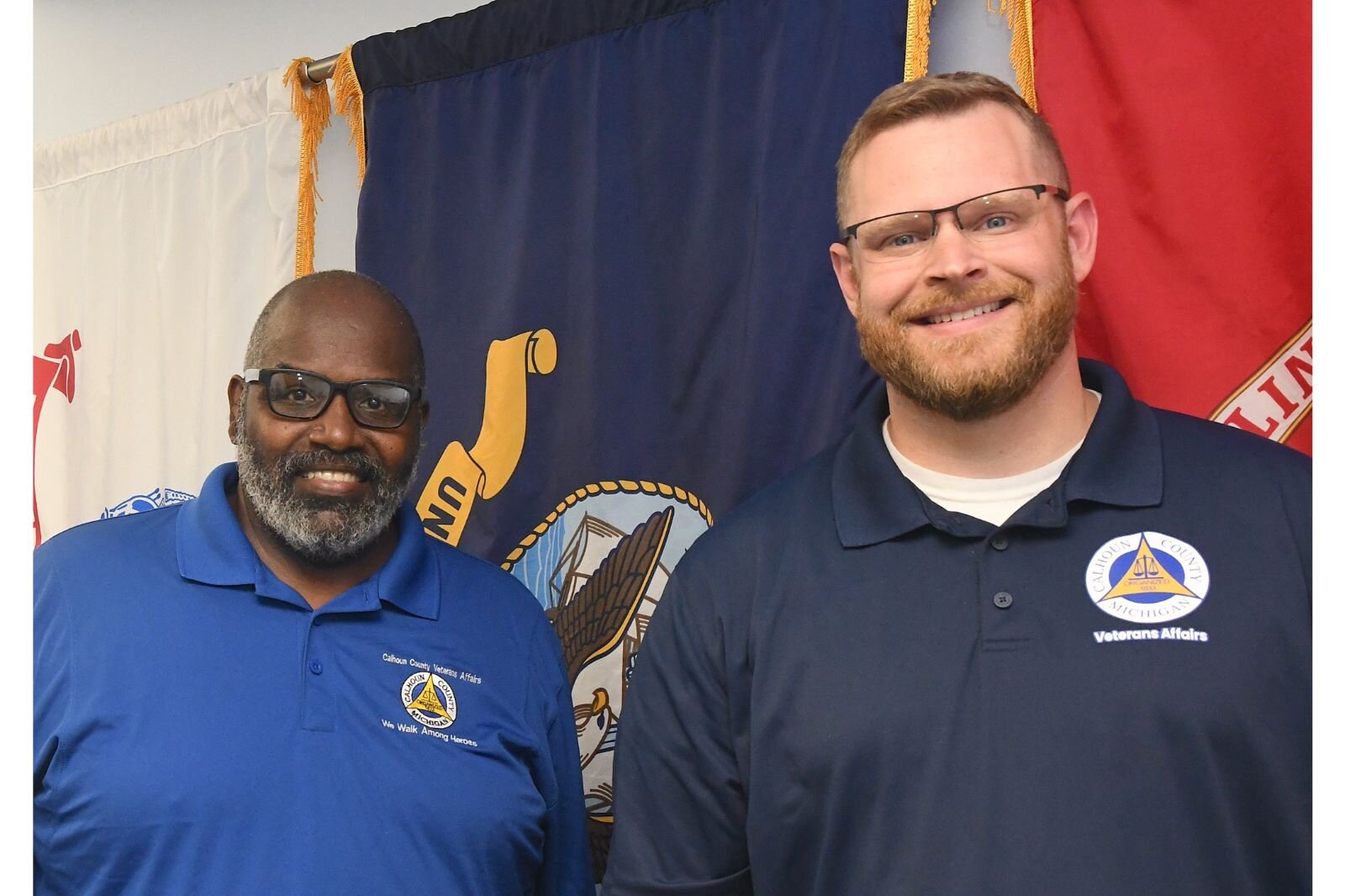 Sam Gray, Chairperson of the Advisory Committee, and Aaron Edlefson, Director, Calhoun County Veterans Affairs, stand in front of military service flags in the organization’s board room.