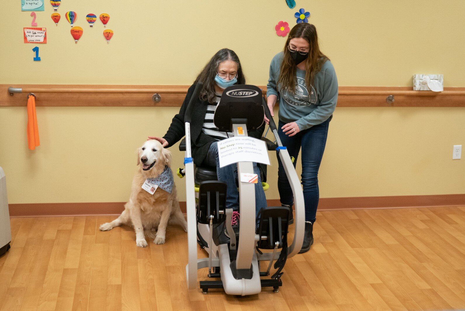 A therapy dog and a staff member help a participant on an exercise bike. 