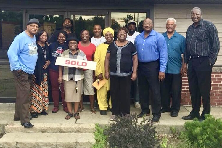 NACD Executive Director Mattie Jordan-Woods holds a “sold” sign as members of the association’s board of directors and the leadership of Faith Temple Church stand in front of a building at the corner of Park and North street in the summer of 2019. 