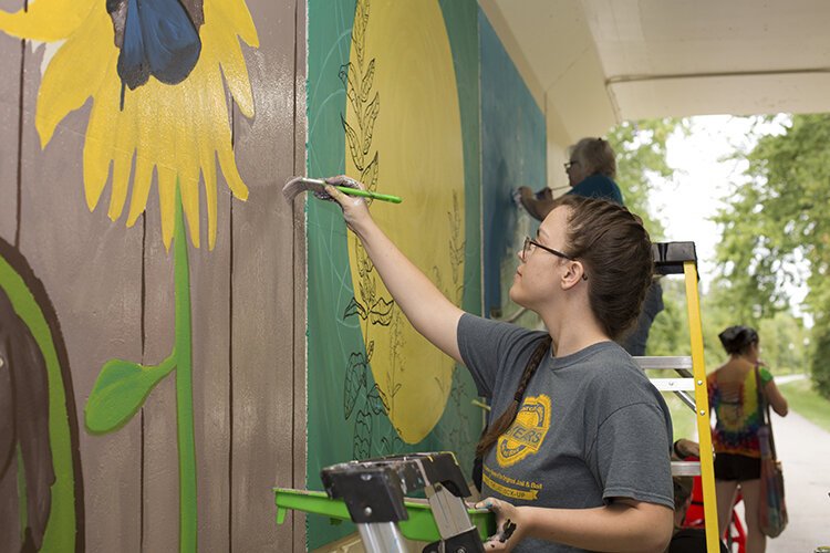 Artist Skylar Todd works on her mural which features sunflowers in front of a fence.